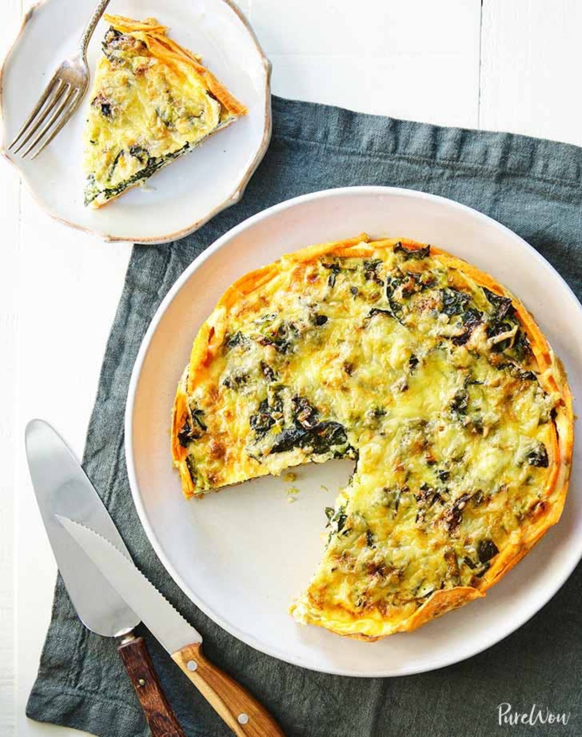 On a white countertop is a square blue napkin. On this is a round white plate containing a quiche with one slice cut out. A cake slice and knife with wooden handles sit below the plate. Above, adnn off of the napkin is a smaller white plate with a portion of quiche on it