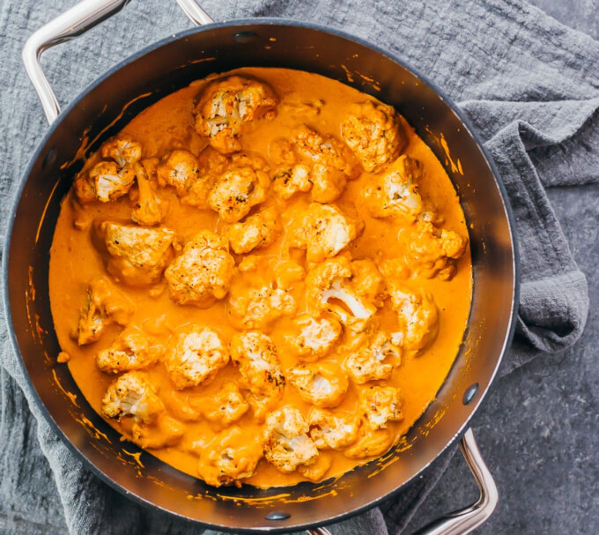 On a rumpled dark blue cloth is a cast iron dish with cauliflower cooked in an orange sauce.