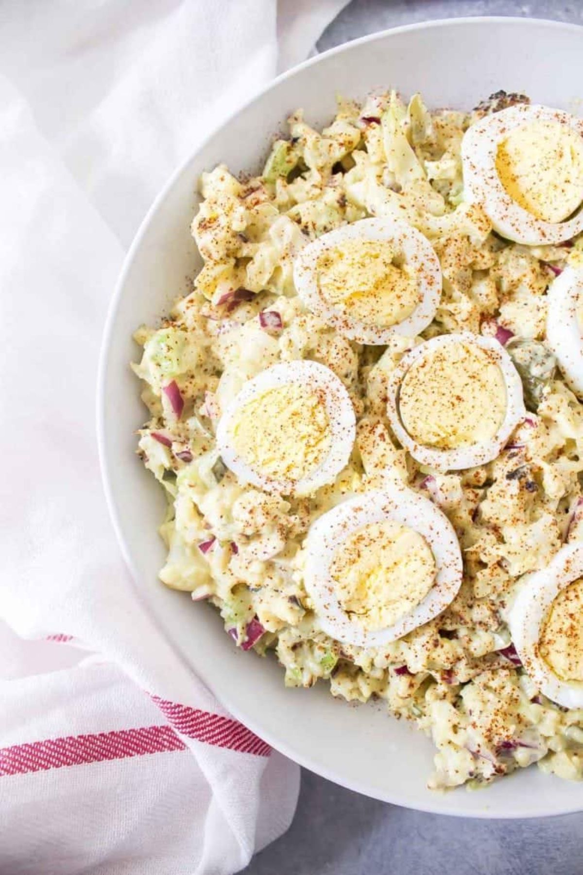 We see half od a white bowl filled with a potato salad, topped with halved boiled eggs and paprika. To the left of the shot is a white cloth with a red stripe at the bottom