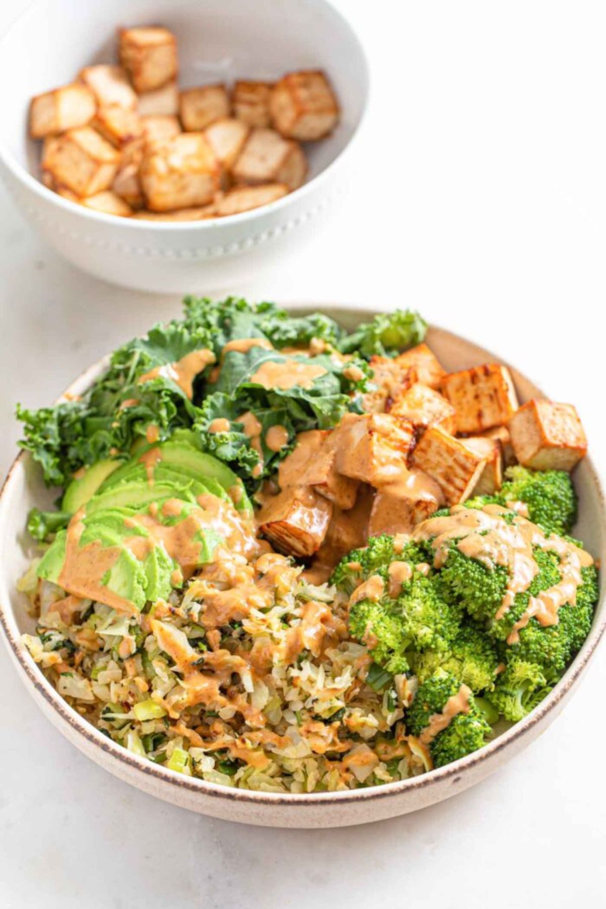 In the foreground is a white bowl with a dark stripe around the rim. Inside are sections of broccoli, fried tofu, kale, avocado and rice, all toppped with an orange sauce. Behind and blurred is a smaller white bowl filled with the tofu