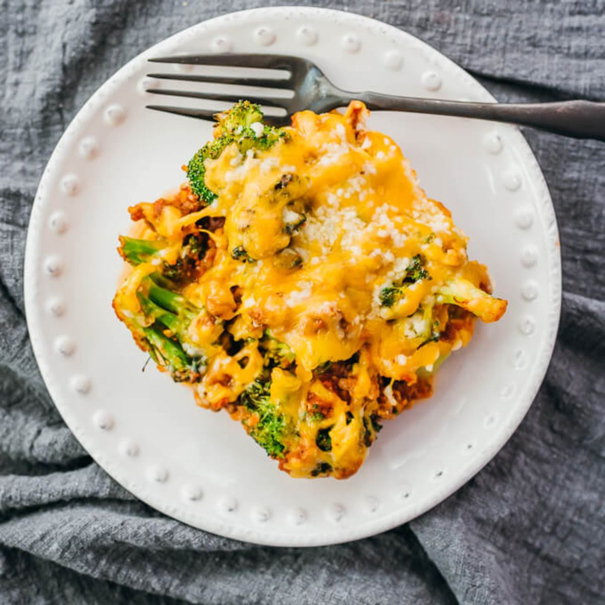 on a white plate is a square portion of casserole with broccoli seen covered in cheese sauce. a metal fork is at the top of the pate