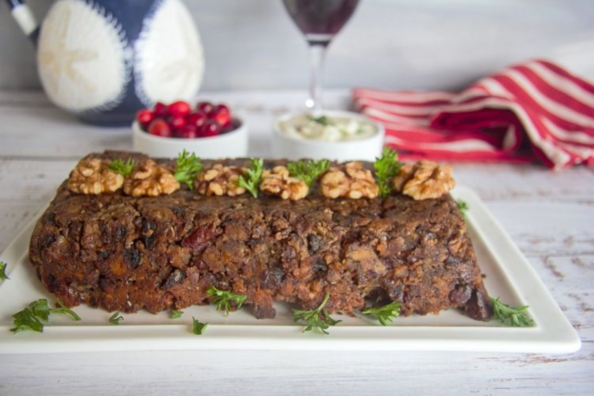 On a whitewashed table is a rectangular board with a wwalnut loaf on top, topped with shelled walnuts and parsley. Behind and blurred are two bowls of cranberries and a white sauce, a glass of red wine, a patterned jug, and a red and white striped towel