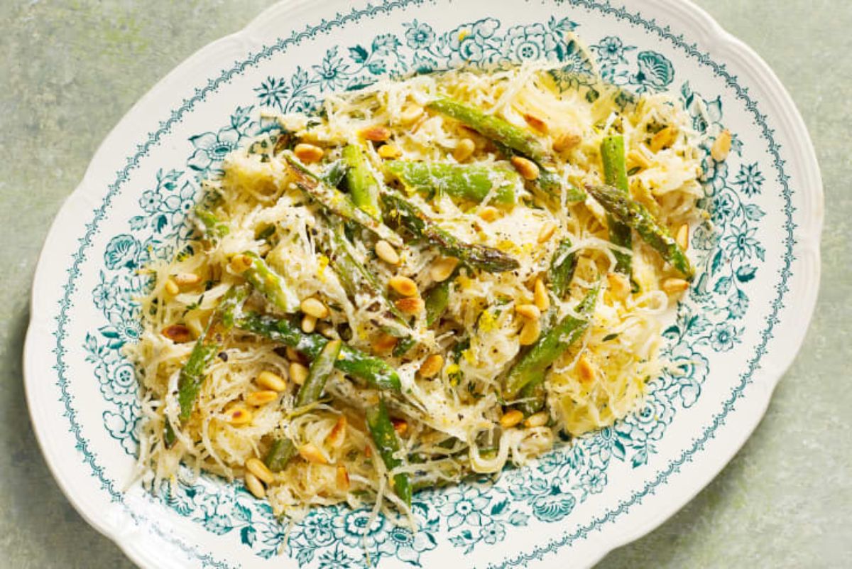 On a cream surface is a patterened charger filled with spaghetti squash and asparagus and topped with pine nuts