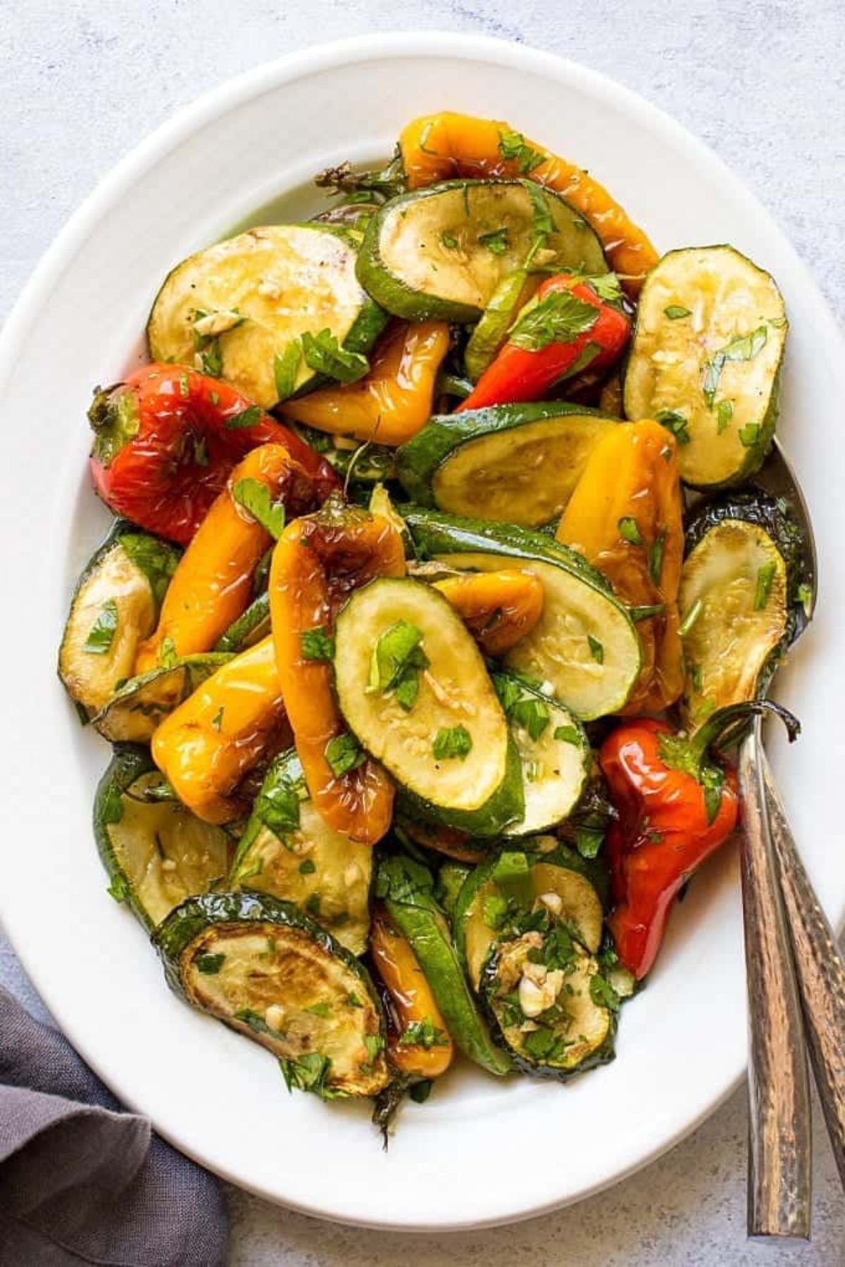 An oval white plate sits on a speckled white surface. On the plate are rosted vegetables including peppers and zucchini. A serving spoon sits on the right.