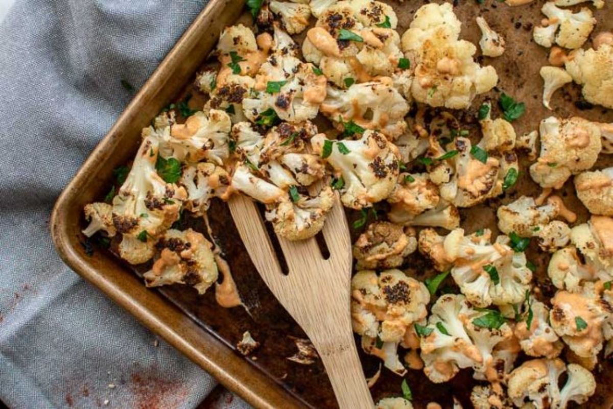 a partial shot of a square shallow roasting dish filled with cauliflower and chopped herbs. It sits on a gray cloth. A wooden fork is stuck into the dish