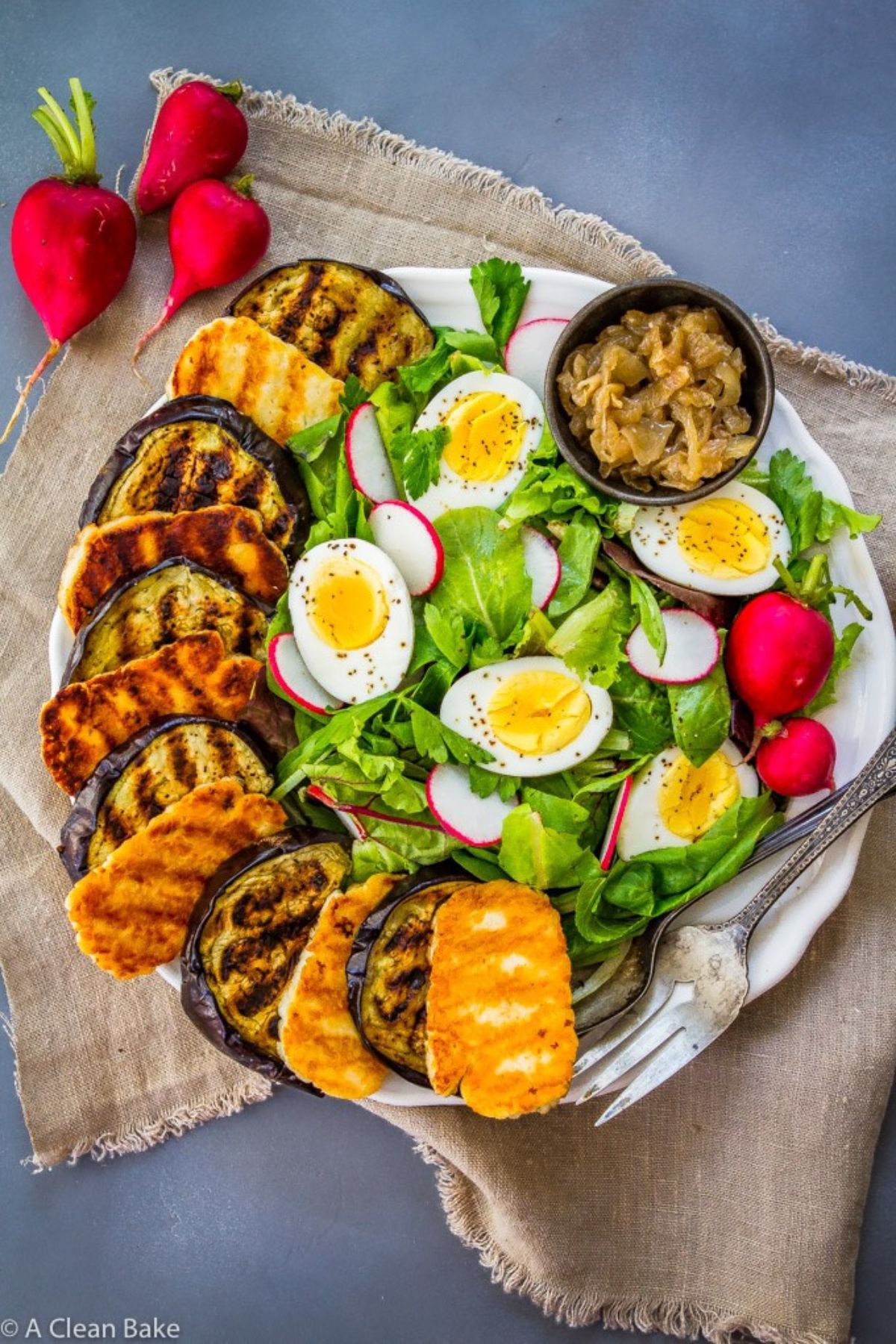 ON a blue counter is a hessian cloth. On top of this is a white round plate with layers of fried eggplant and halloumi on the left, and salad leaves radishes and halved boiled eggs on the right. A small pot of pickle sits at the top of the plate. To the left are 3 radishes