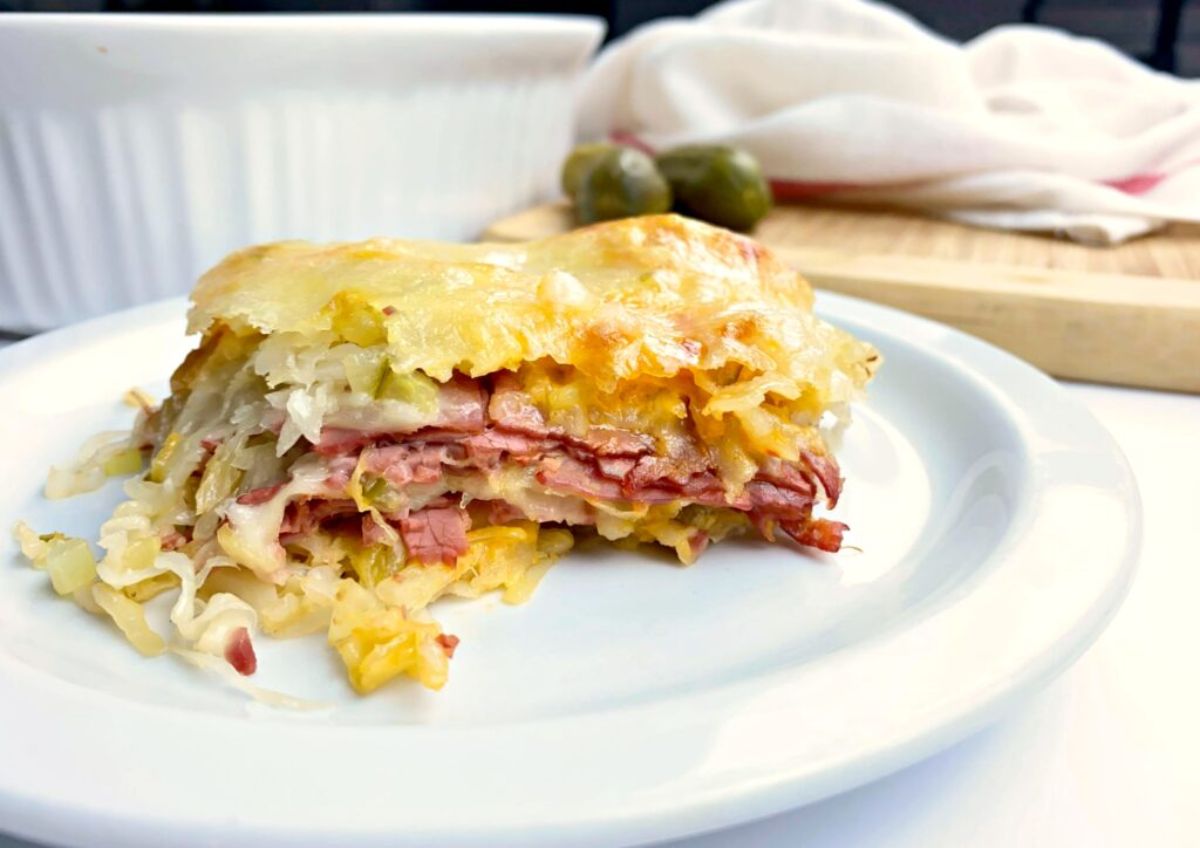 in th background a blurred wooden chopping board and a white towel on top. In the foreground a white plate with a square portion of layered cassserole includng meat and cabbage