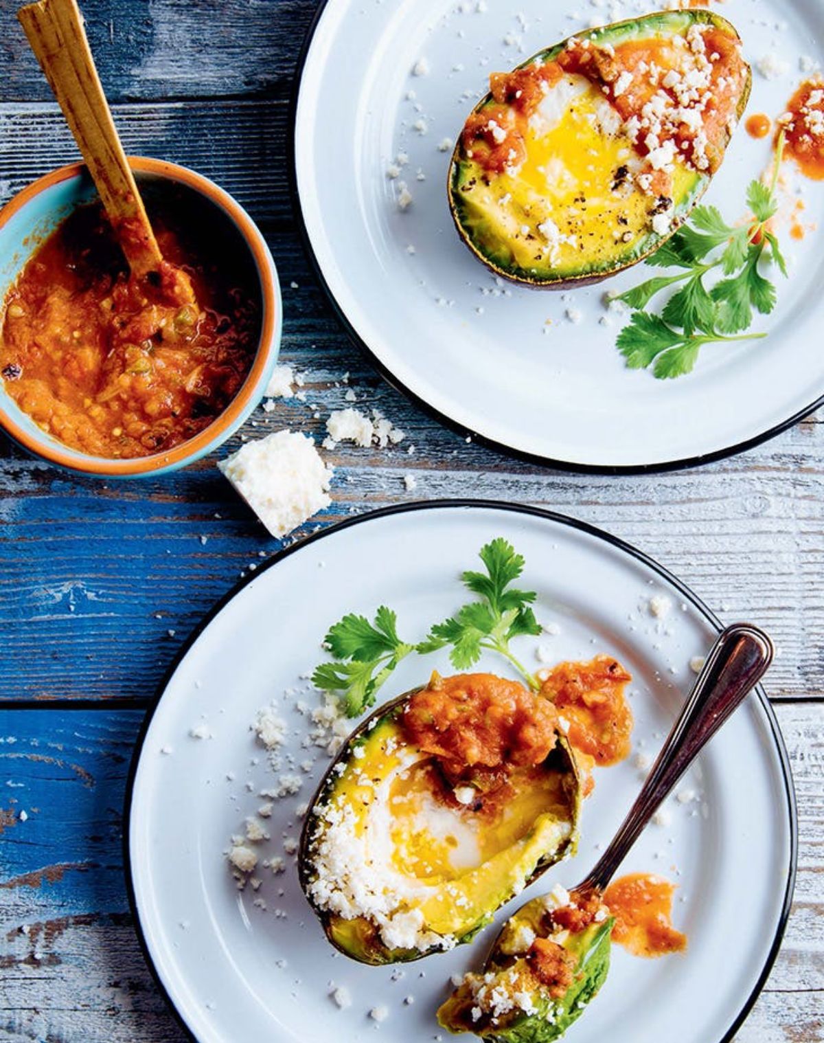 On a painted blue table are 2 white plates with black rims. On each is half an avocado with a baked egg inside it, parmesan sprinkled over the top and parsely sprigs on the side. On the left is a blue pot with red sauce in it and a small wooden spoon