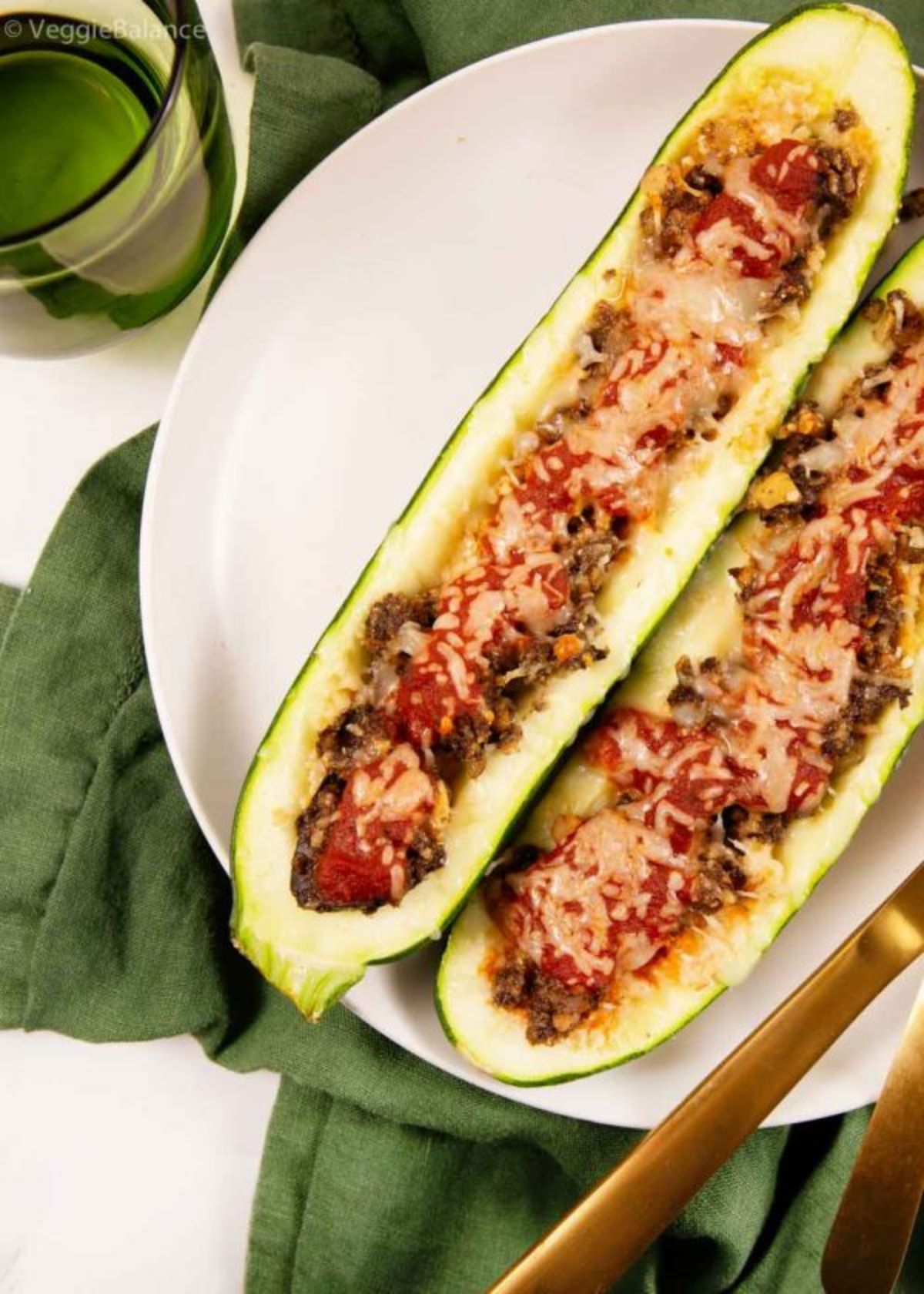 On a green cloth is a white round plate. On this are two zucchini halves stuffed with a tomato and cheese mixture. THe handles of golden cutlery can be seen to the bottom right and a glass to the top left