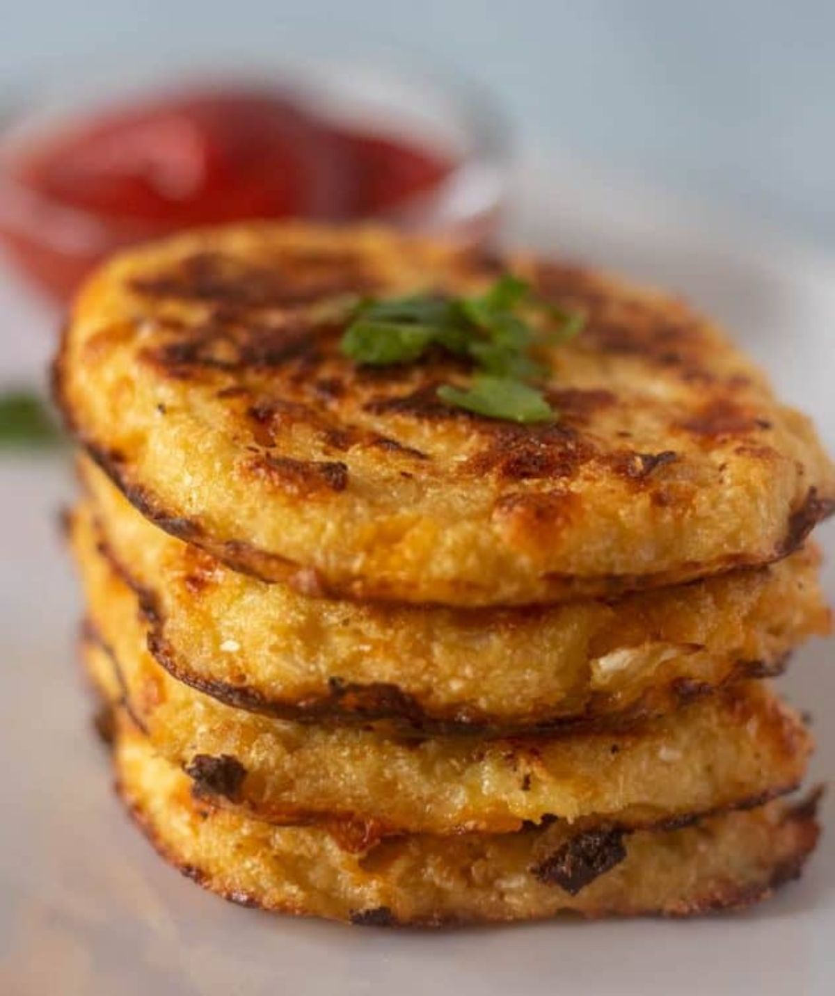 WIth a blurred background of a pot of red sauce, a stack of four hash browns with chopped herbs on top