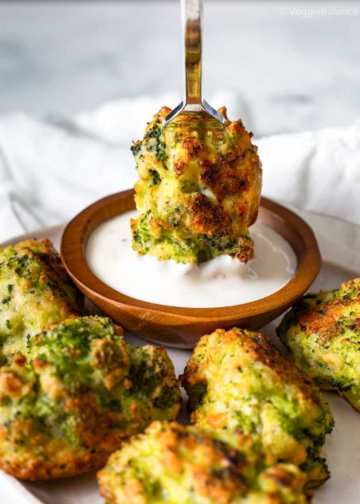 On a white background is a whote round plate with baked broccoli florets. In the middle is a bronze smal bowl with white sauce in it. A broccoli floret is being dipped in to the sauce using tongs