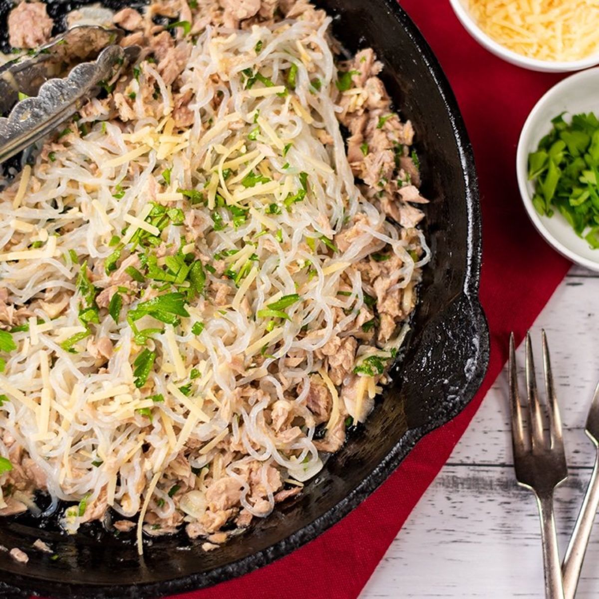 a partial shot of a white wooden tablewith a red cloth over part of it. On the cloth is a cast iron pan with tuna spaghetti in it. To the right are bowls of chopped herbs and grated cheese