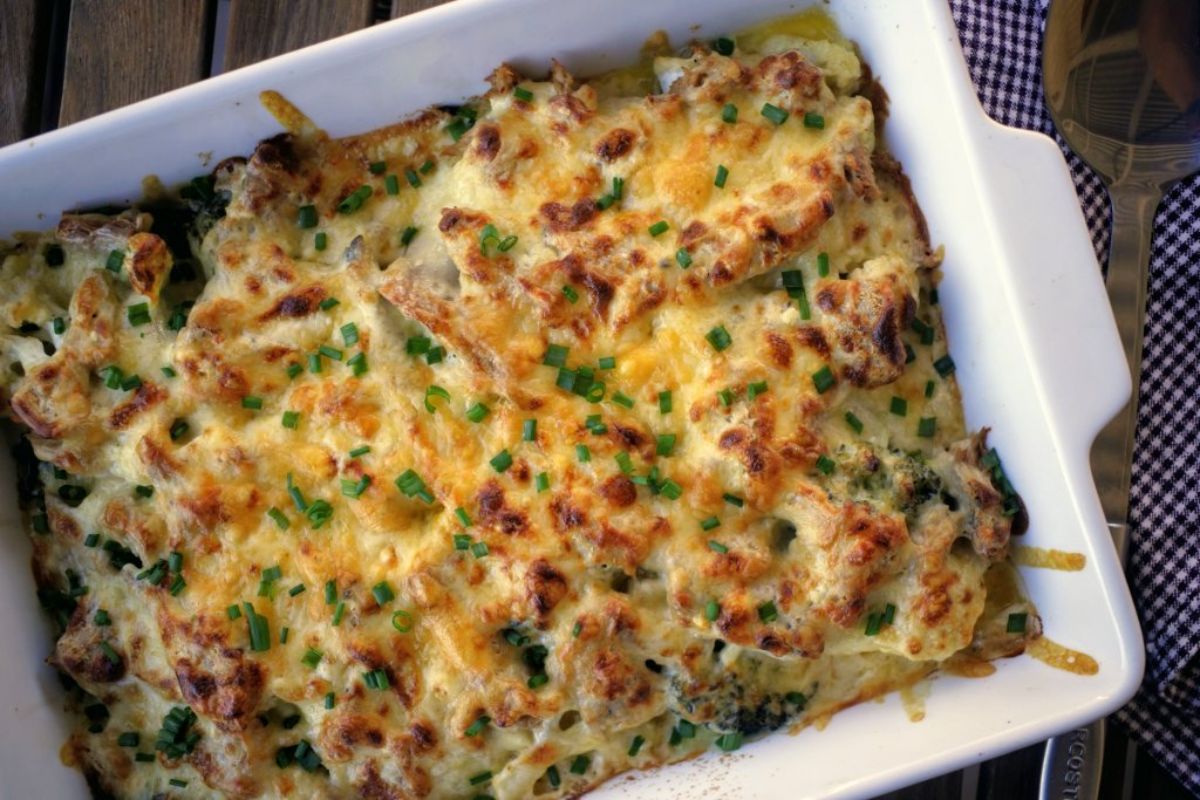 A partial shot of a wooden table with a checked sheet and a wooden serving spoon. The rectangular casserole dish is filled with cheesy casserole and topped with chopped herbs