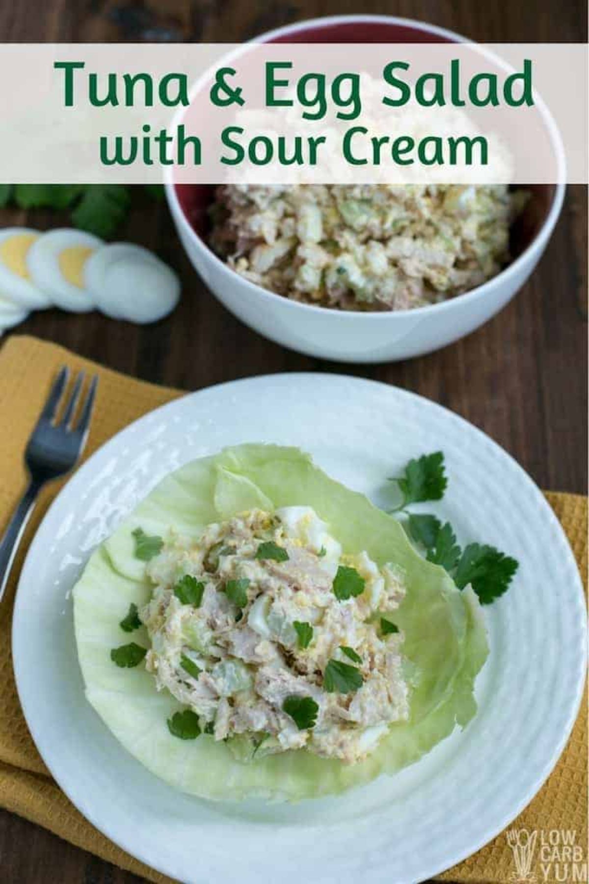 The text reads "Tuna & Egg salad with sour cream". The photo is of a wooden table with a yellow rectangular cloth with a white round plate on top of it. A lettuce leaf is filled with tuna and egg salald and sprinked with herbs. Behind is a deep white bowl with the salad in it. To the left are some slices of boiled egg and a silver fork