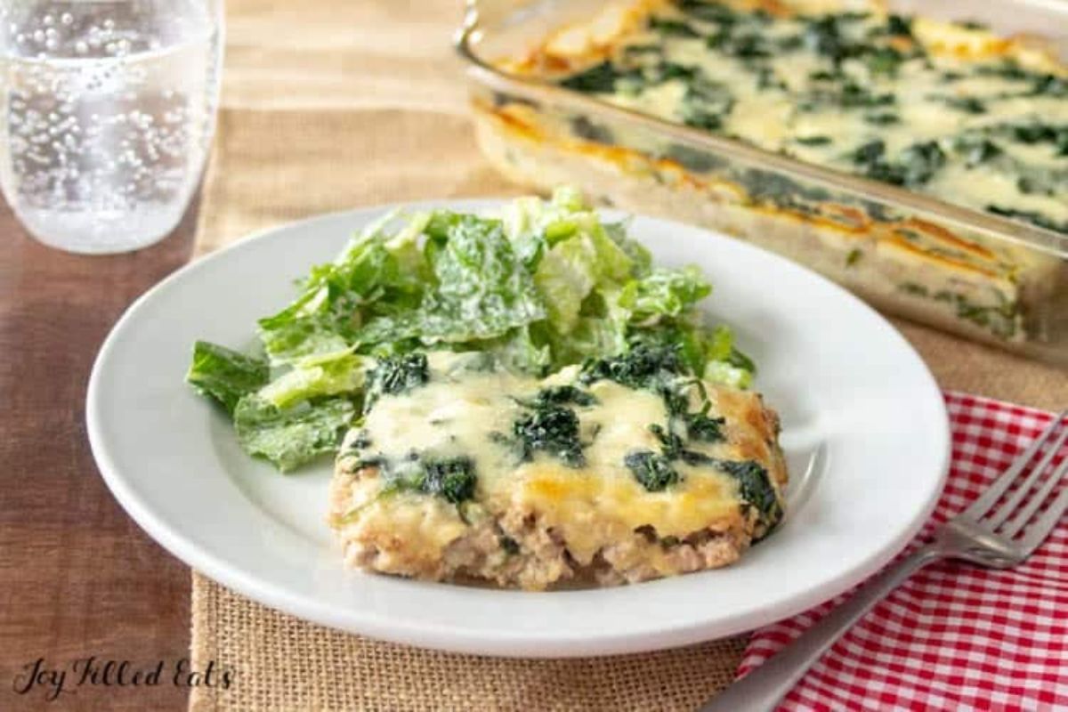 On a wooden table, surrounded by a glass of water, and a lurred shot of a rectangular casserole dish filled with creamy spinach casserole, is a square portion of the casserole garnished with a side of green lettuce