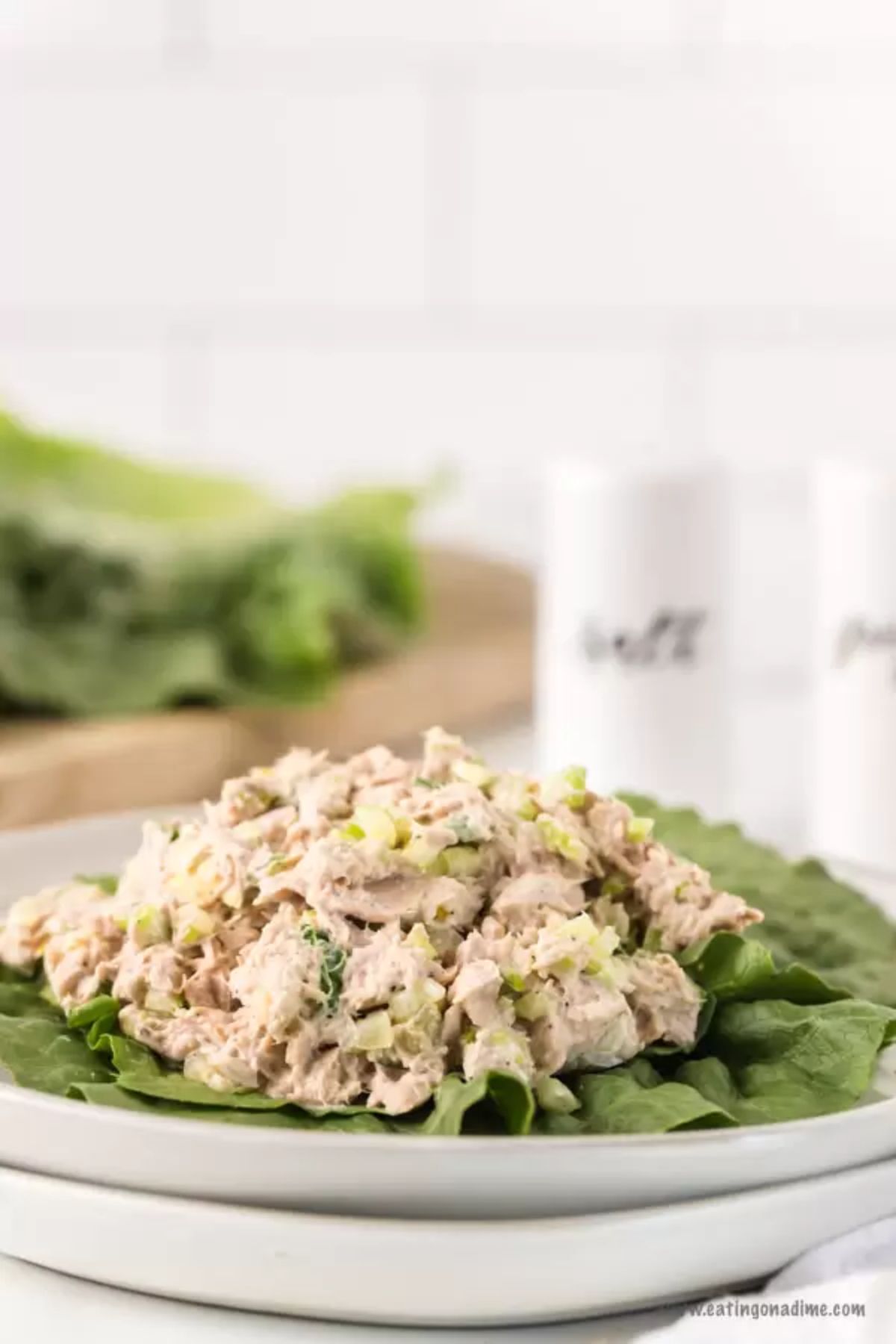 In the foreground is a stack of two white plates with lettuce leaves piled with tuna salad, Blurred in the background are some lettuce leaves on a wooden board and white salt and pepper shakers