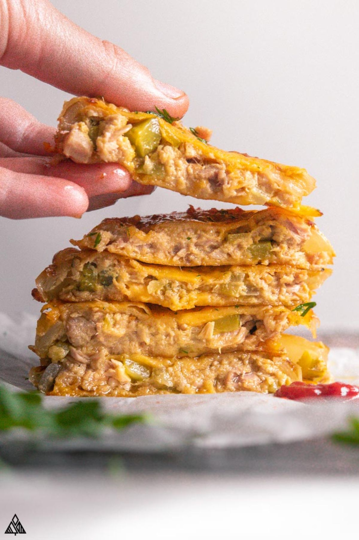 A hand reaches in from the left to pick up the top patty from a stack of 5 filled with cheesy tuna.