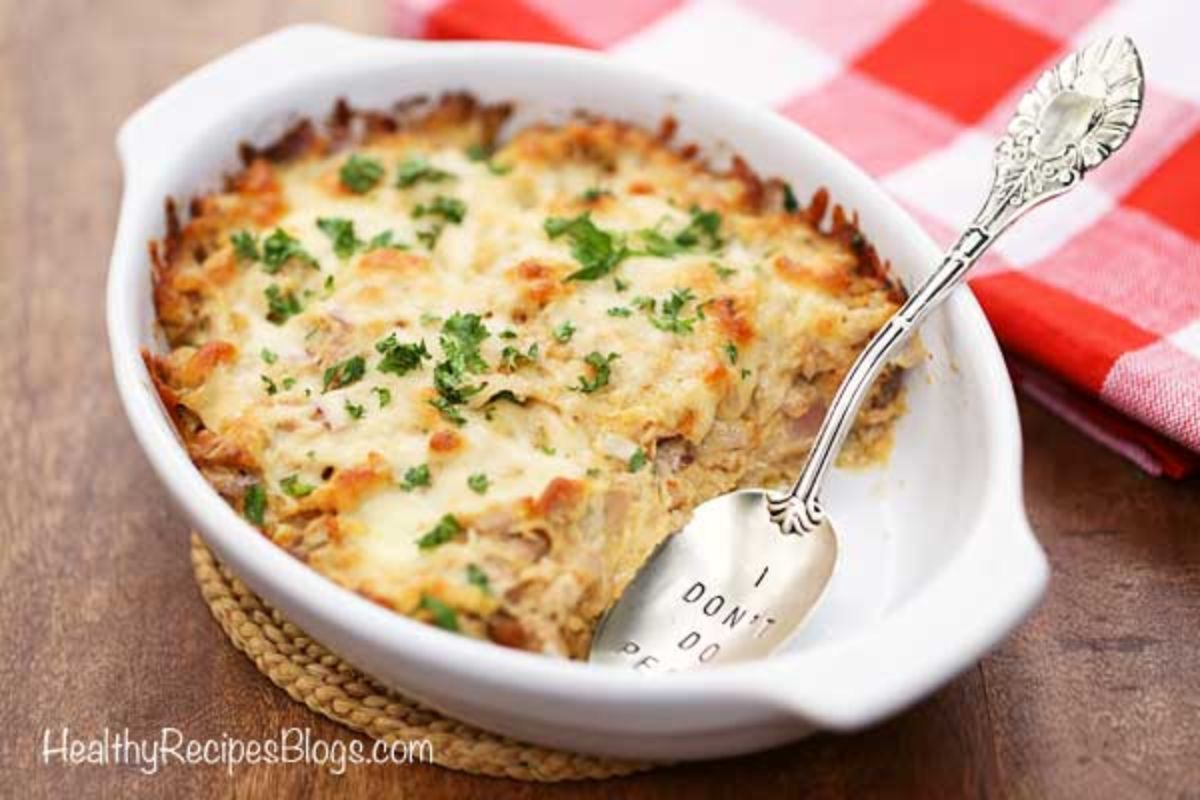 On a dark wooden table is an oval white cassreole dish filled with casserole and topped with chopped herbs. A silver spoon rests in the front of the dish. A red and white cloth is behind