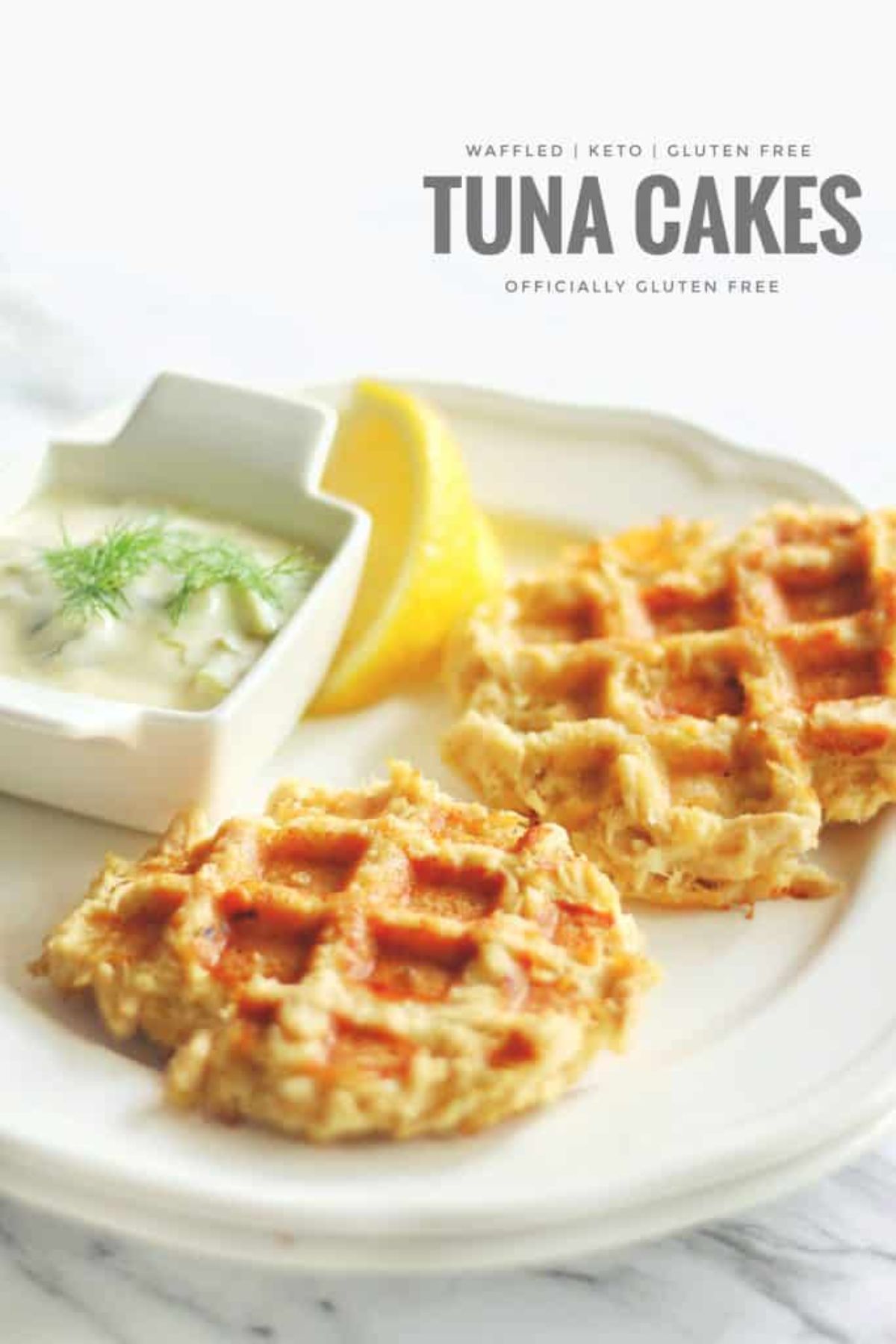 The text reads "Waffled, Keto, Gluten-free Tuna cakes. Officially gluten-free" On a white marbled surface is a white plate with two waffles, a lemon slice and a square pot full of white sauce