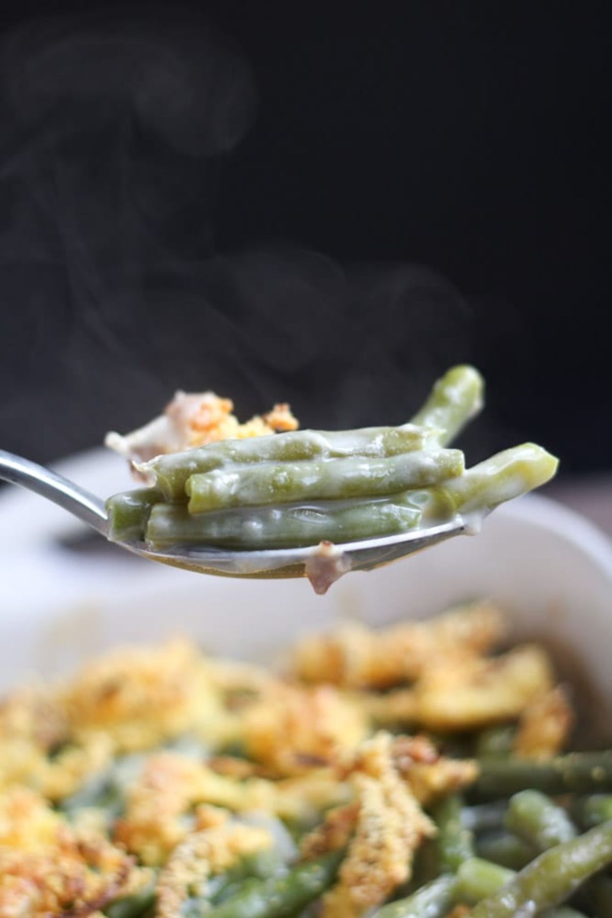 In the background is a white dish of green bean casserole. In the foreground a metal spoon with a spoonfull of the green beans on it