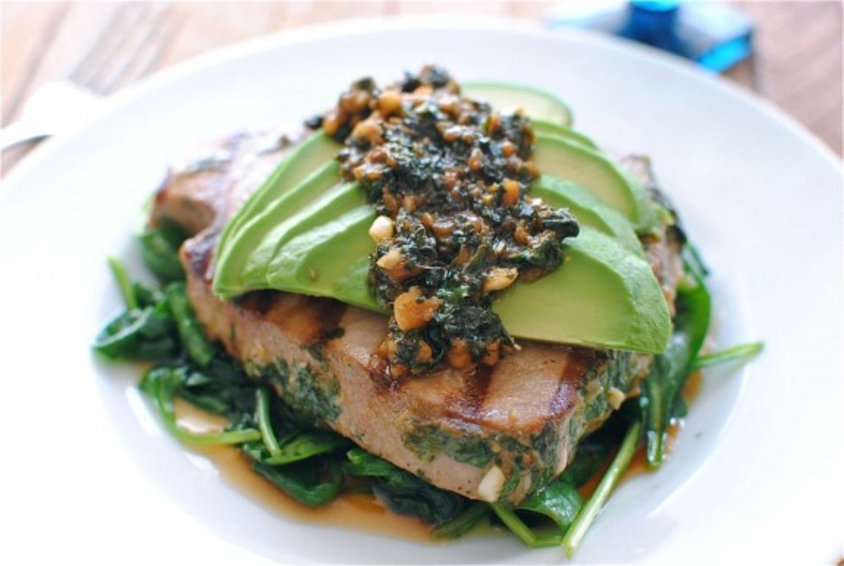 A white round plate with wilted spinach, seared tuna steak, sliced avocado and pesto