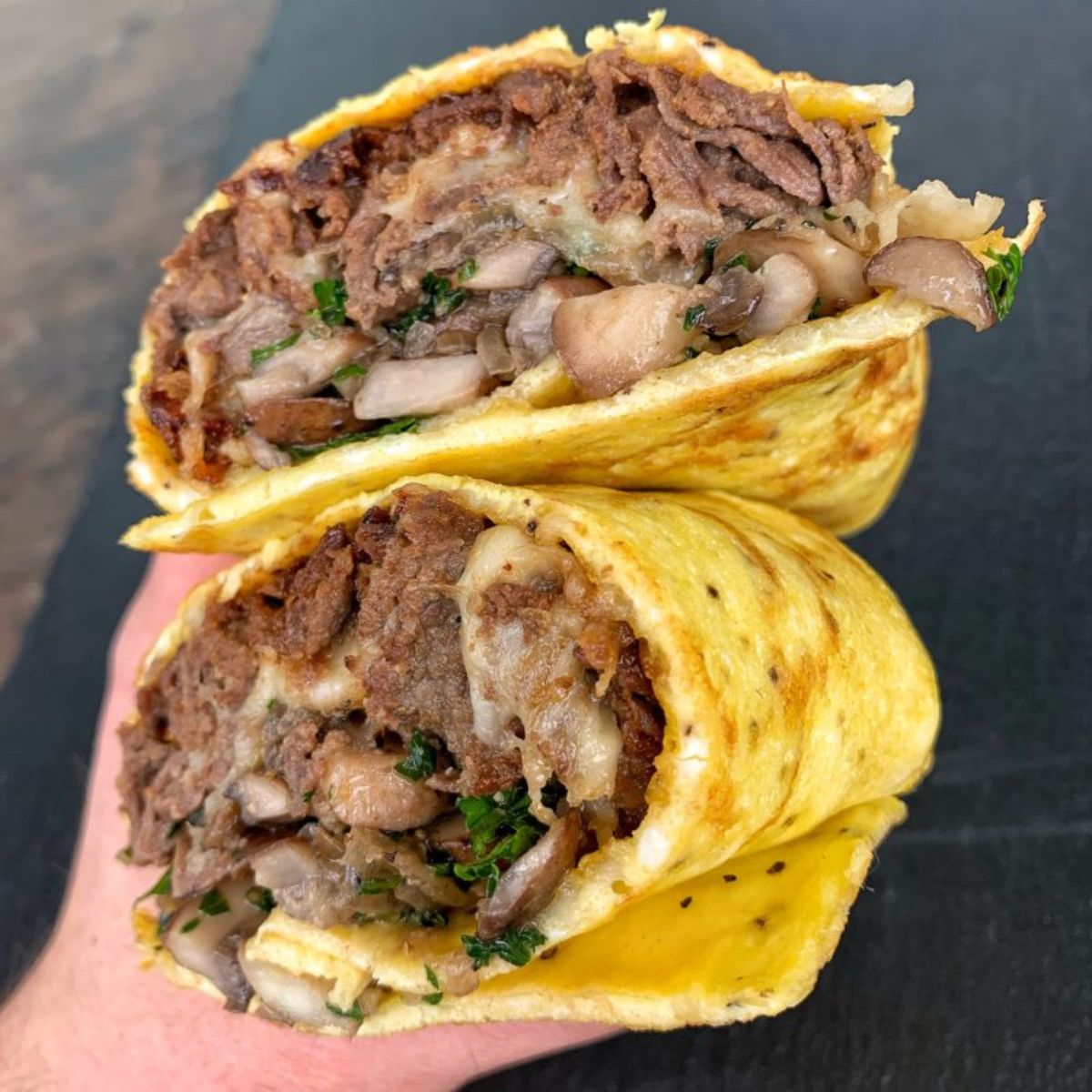 A hand holds two halves of an egg burrito filled with steak and vegetable mix