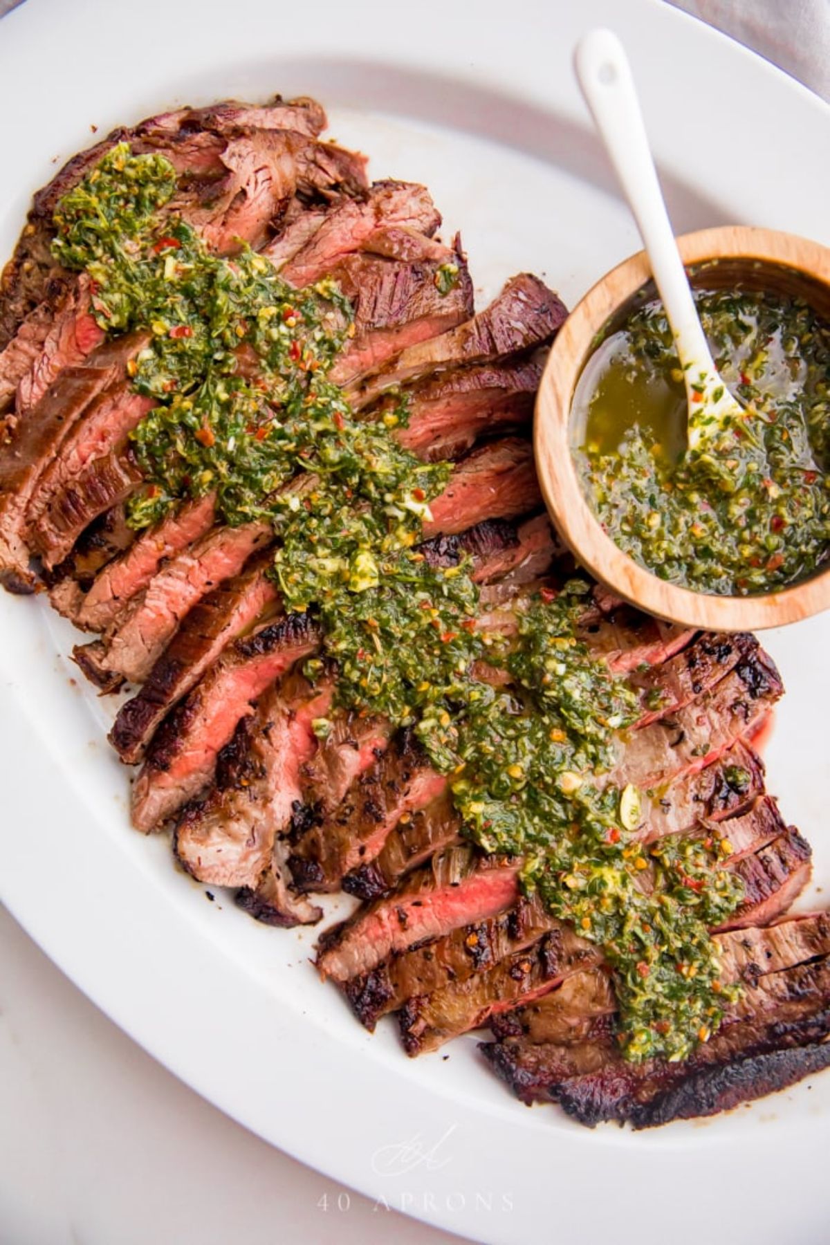 An oval white plate holds slices of rare steak, drizzled over with chiichurri sauce. A wooden pot of the sauce sits on the right of the plate with a white spoon in it