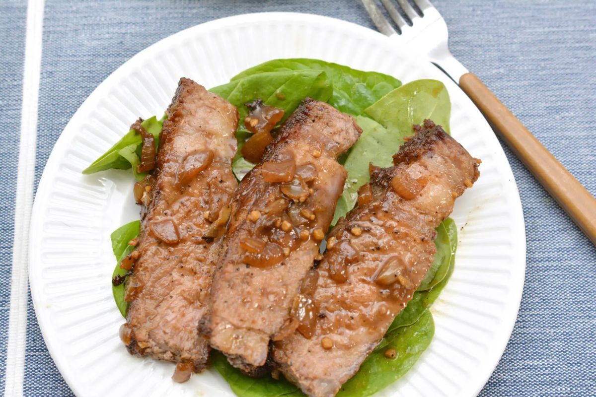 A round white plate sits on a blue cloth. On the plate is a bed of leaves wth 3 clices of steak on top. To the right of the plate is a wooden handled fork