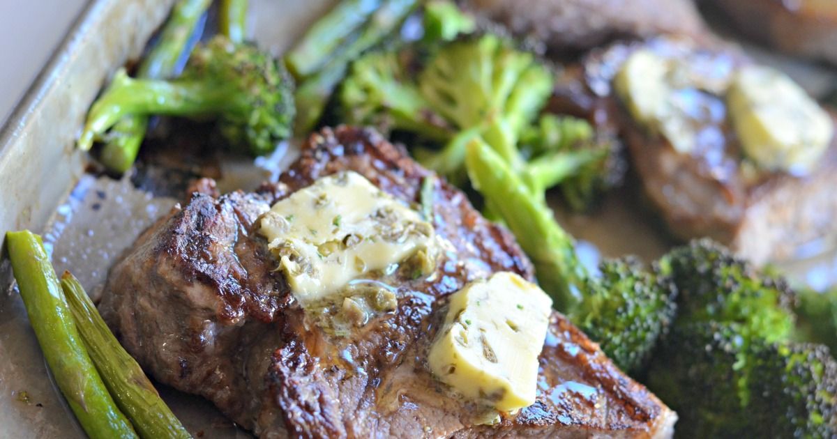 A partial shot of a baking tray filled with broccoli florets and steak. On the steak are two slabs of garlic butter