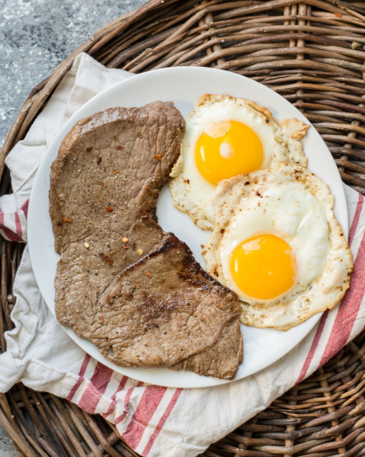 On a wicker tray is a white and red cloth, on which rests a white plate. On the plate is a large steak and 2 fried eggs.