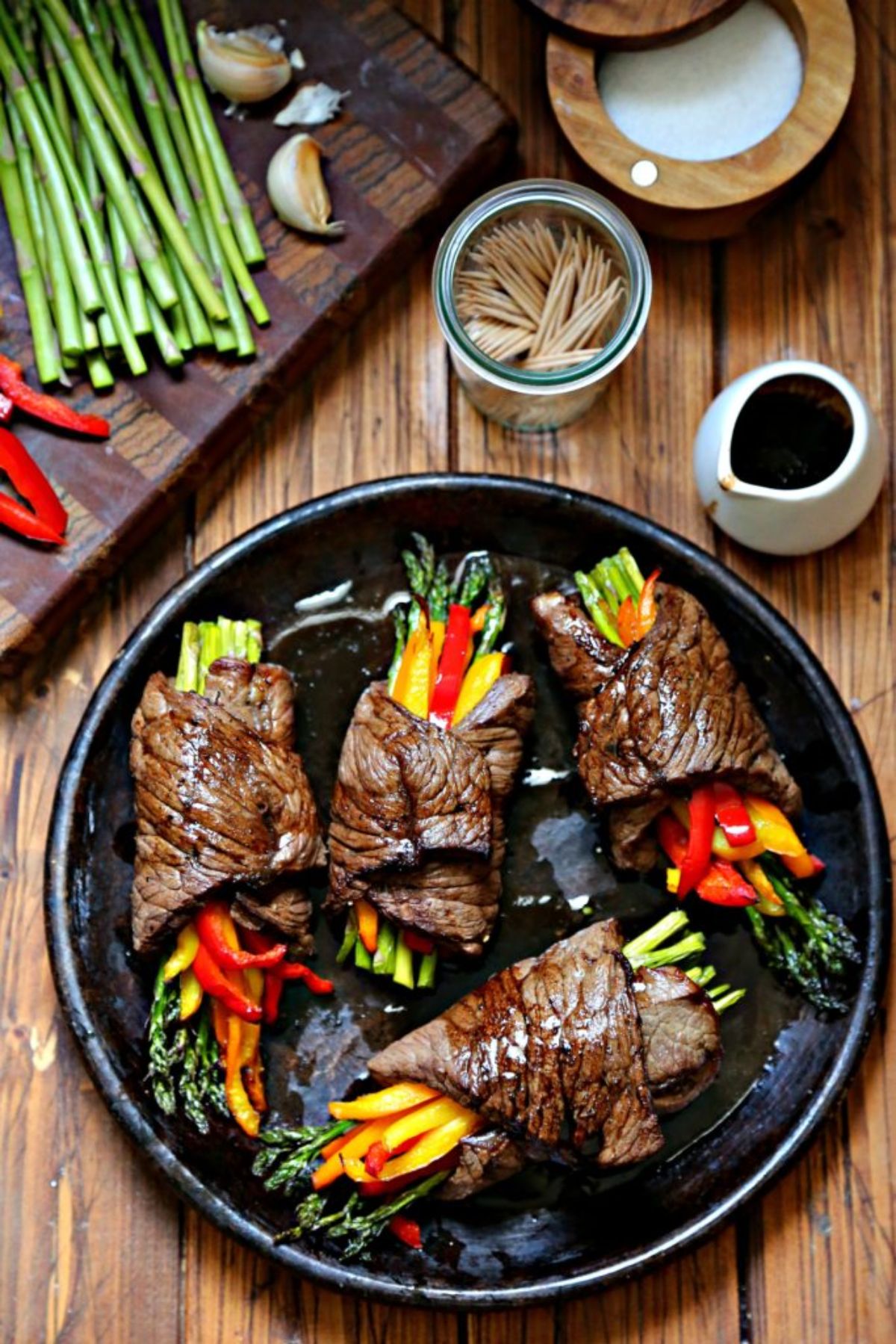 A dark wooden tale has a chopping board on the top left filled with asparagus, sliced peppers and garlic cloves. On the top right is a woodean bowl full of salt, a glass jar full of toothpicks and a white jug full of sauce. At the bottom fo the shot is a dark round plate with 4 bundles of asparagus and pepper wrapped in steak slices.