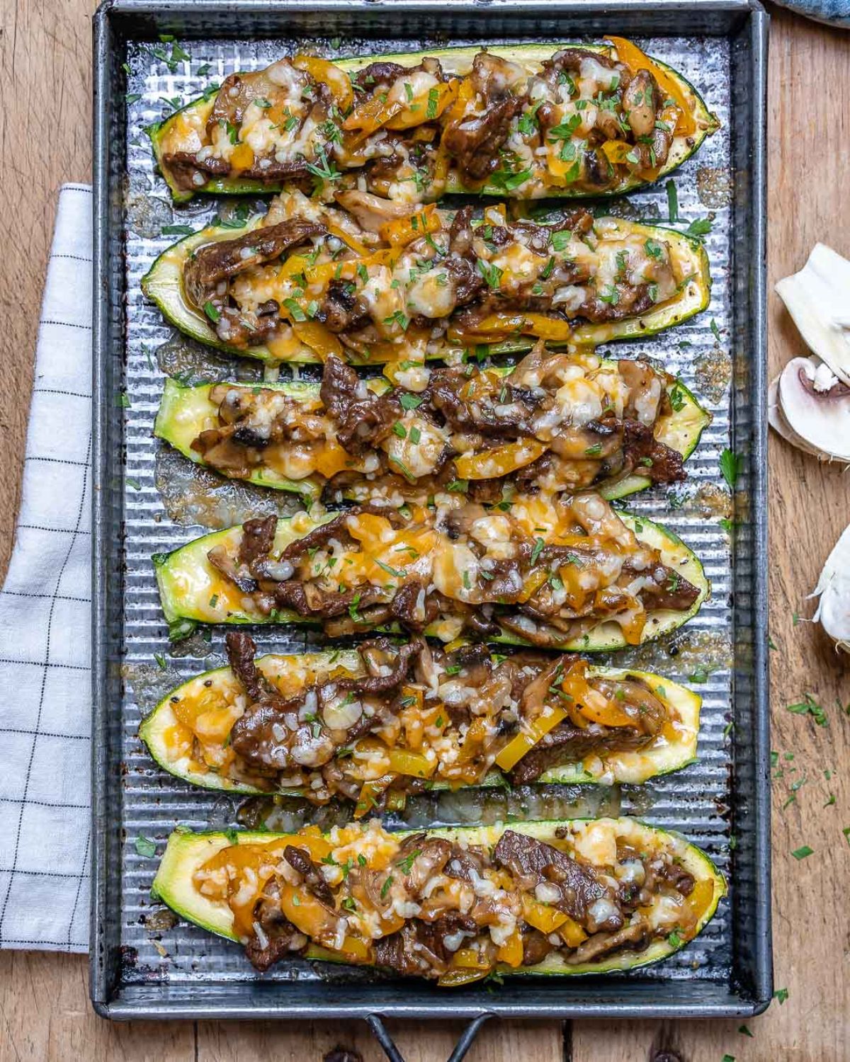 A rectabgular baking sheet sits on a wooden table. 6 zucchini halves are laid out horizontally and filled with philly cheesesteak mixture