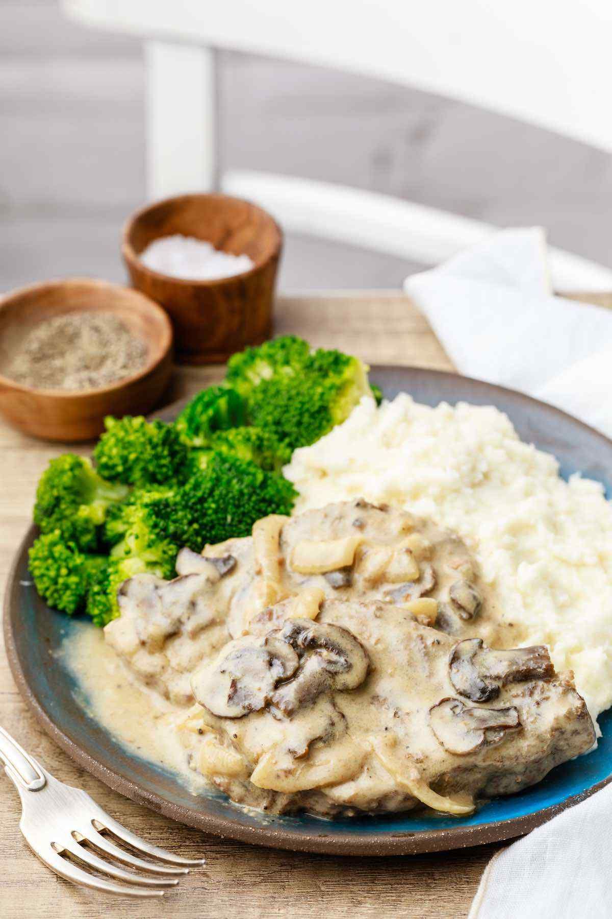 A blurred shot of a table and white chair. On the table in the foreground is a blue plate with broccoli, mash and a steak covered in creamy mushroom sauce.