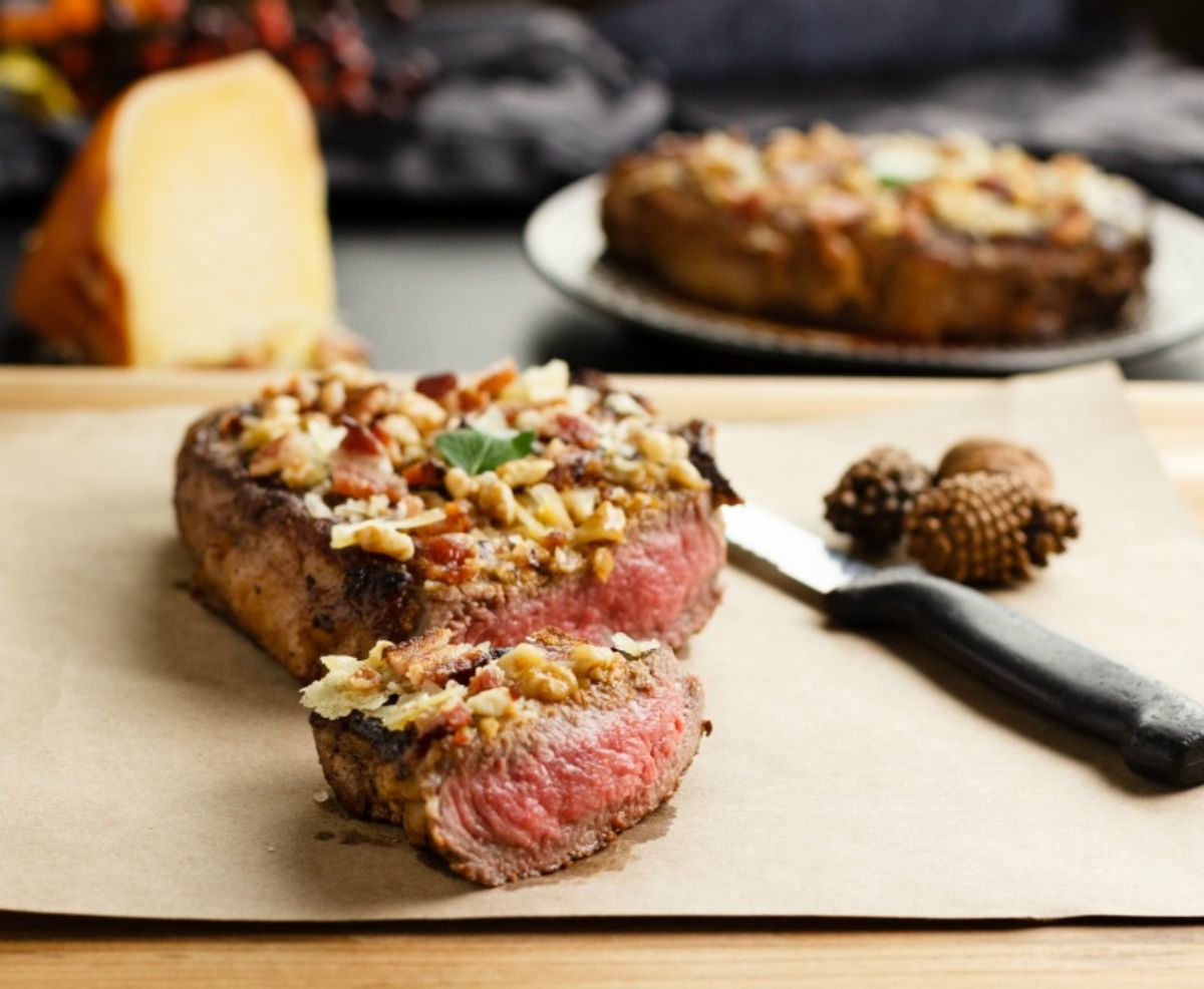 Blurred in the background on a table is a chunk of cheese and a plate with a steak on it. In the foreground on a baking sheet is a thick steak, cut in half and topped with cheese, bacon and walnuts. A steak knife and some pine cones are to the right
