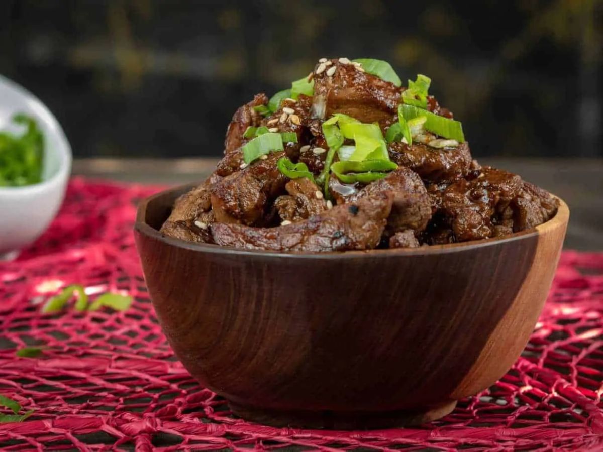 On a red patterened mat is a deep wooden bowl. In it are slices of beef toped with sesame seeds and sliced scallions