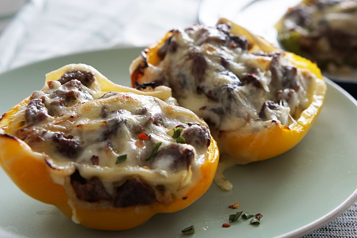 A white, oval plate holds 2 halves of a yellow pepper. Inside the halves is a cheesesteak mixture