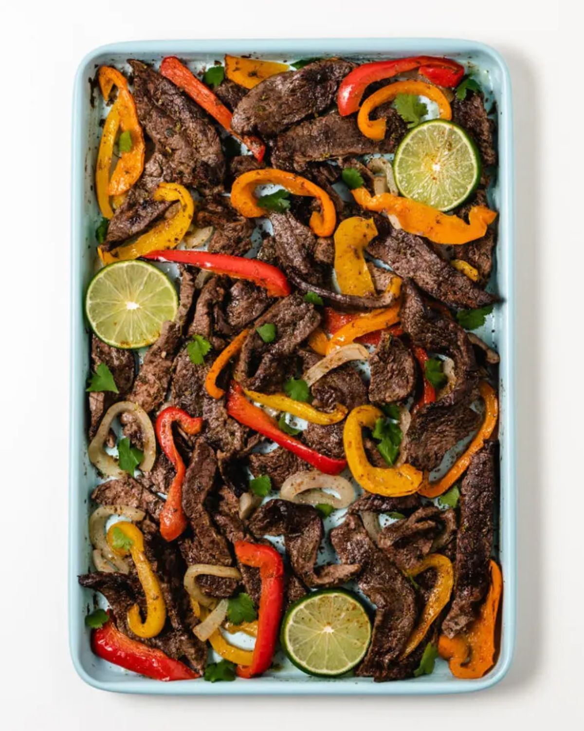 A rectangular roasting dish filled with steak slices, peppers, onions and lime slices.