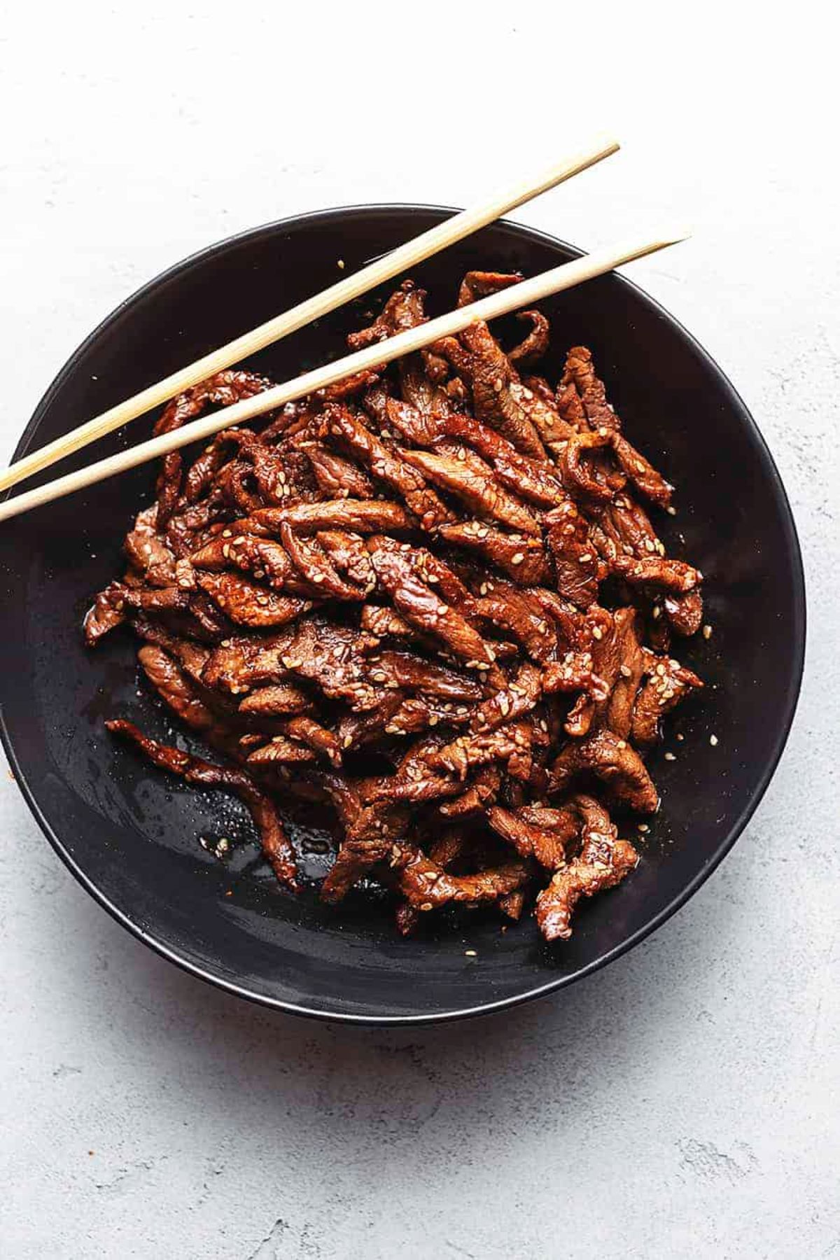 A white background with a black china bowl. Stips of steak in sauce are inside, with 2 chopsticks laid over the top of the bowl