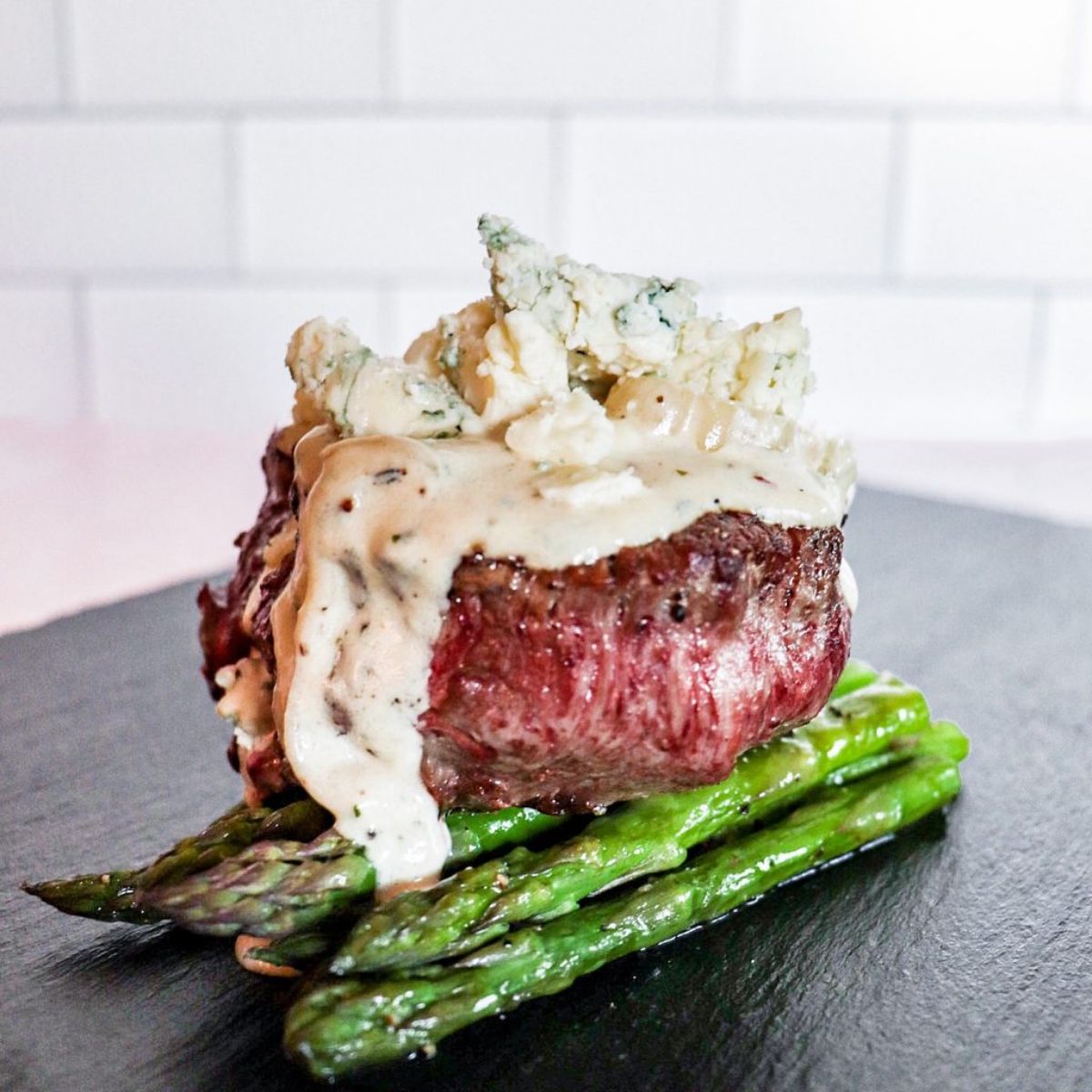 On a dark wooden board against a black background is a bunch of asparagus, with a thick steak on top, topped by a cheese sauce