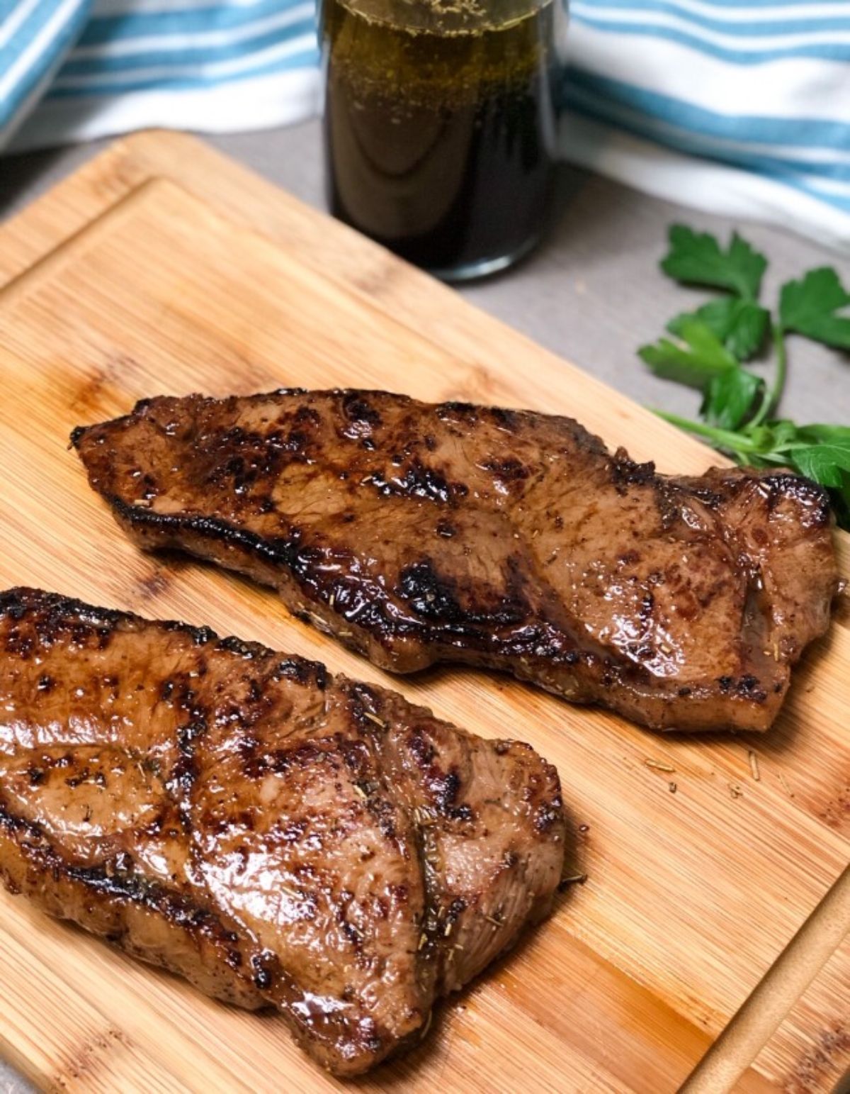 A wooden chopping board is on a table in front of a full glass and some herbs. On the board are 2 cooked flank steaks