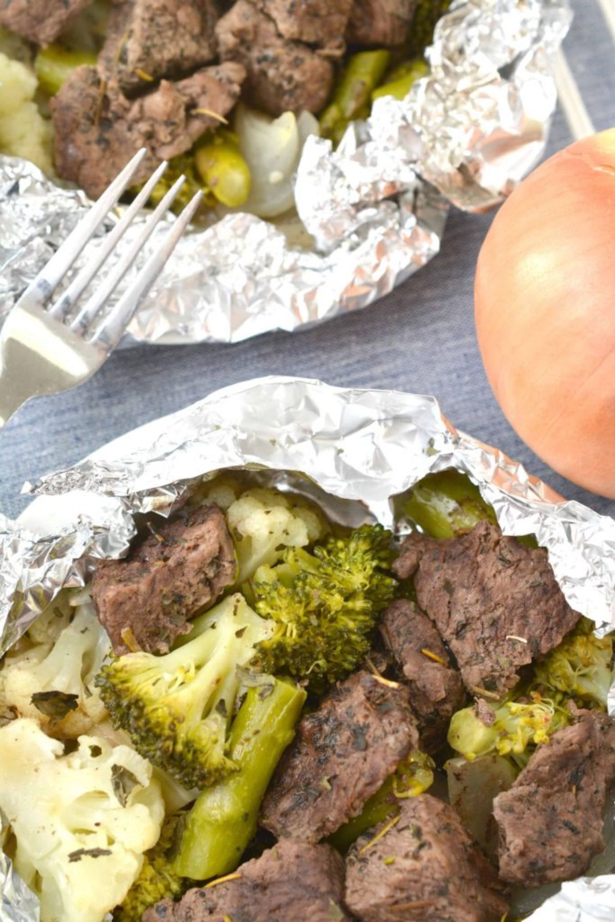 2 foil pockets are on a countertop, slightly undone. Inside are steak bites and broccoli florets. There is the top of a fork digging into one pocket