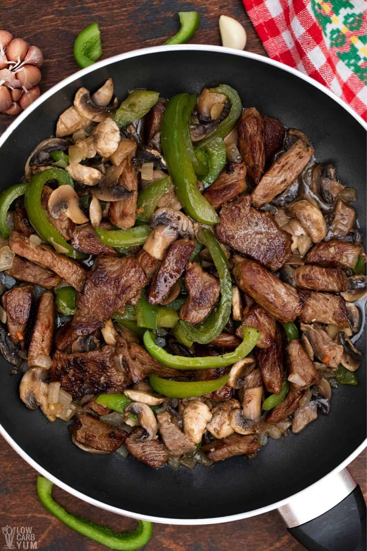 A top view of a skillet filled with steak slices, sliced mushrooms and sliced green peppers