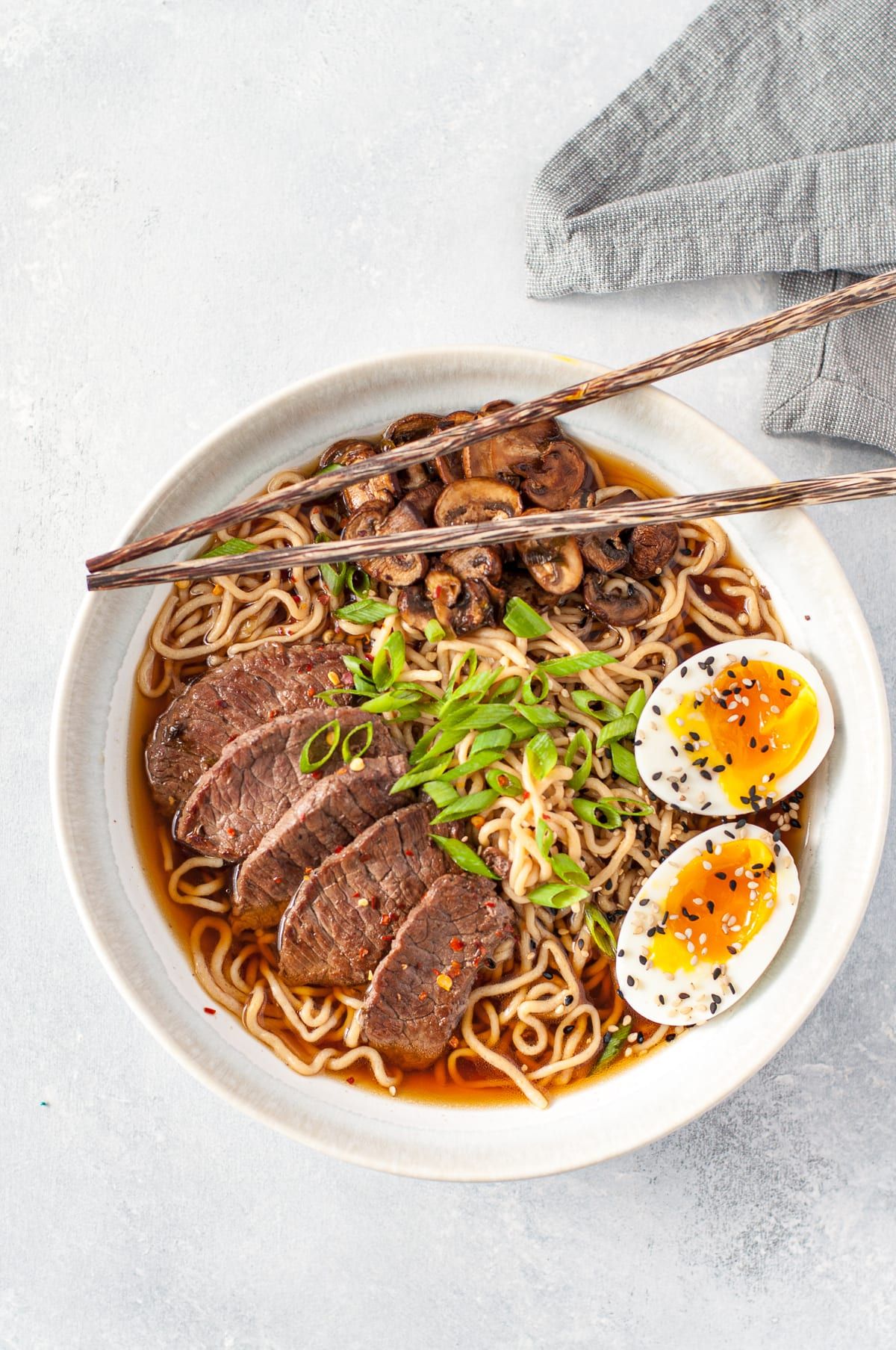 On a white background is a white bowl with ramen noodles, 2 halves of a boiled egg, slices of steak, sliced ushrooms and two chopsticks resting on the top of the bowl. A gray cloth can just be seen top right