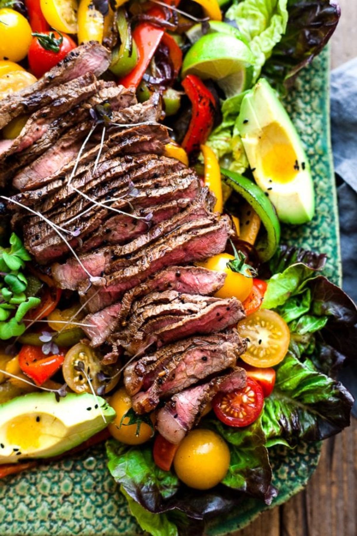 A partial shot of a green serving platter. On it are lettuce leaves, avocado, sliced peppers, halved yellow and red tomatoes, and sliced steak