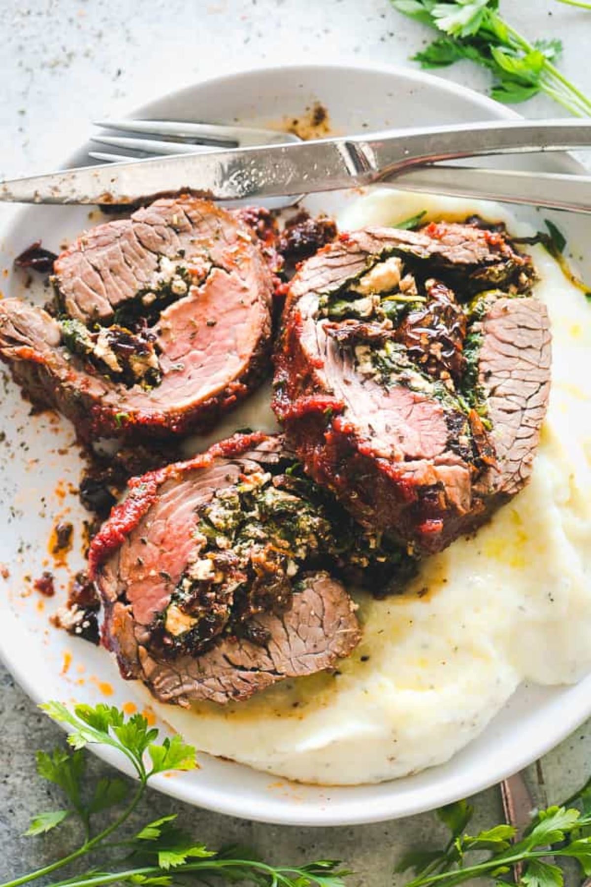 A white plate holds 3 slices of stuffed flank steak on a bed of mash. A knife and fork are at the top of the plate resting on each other. Herbs are scattered around