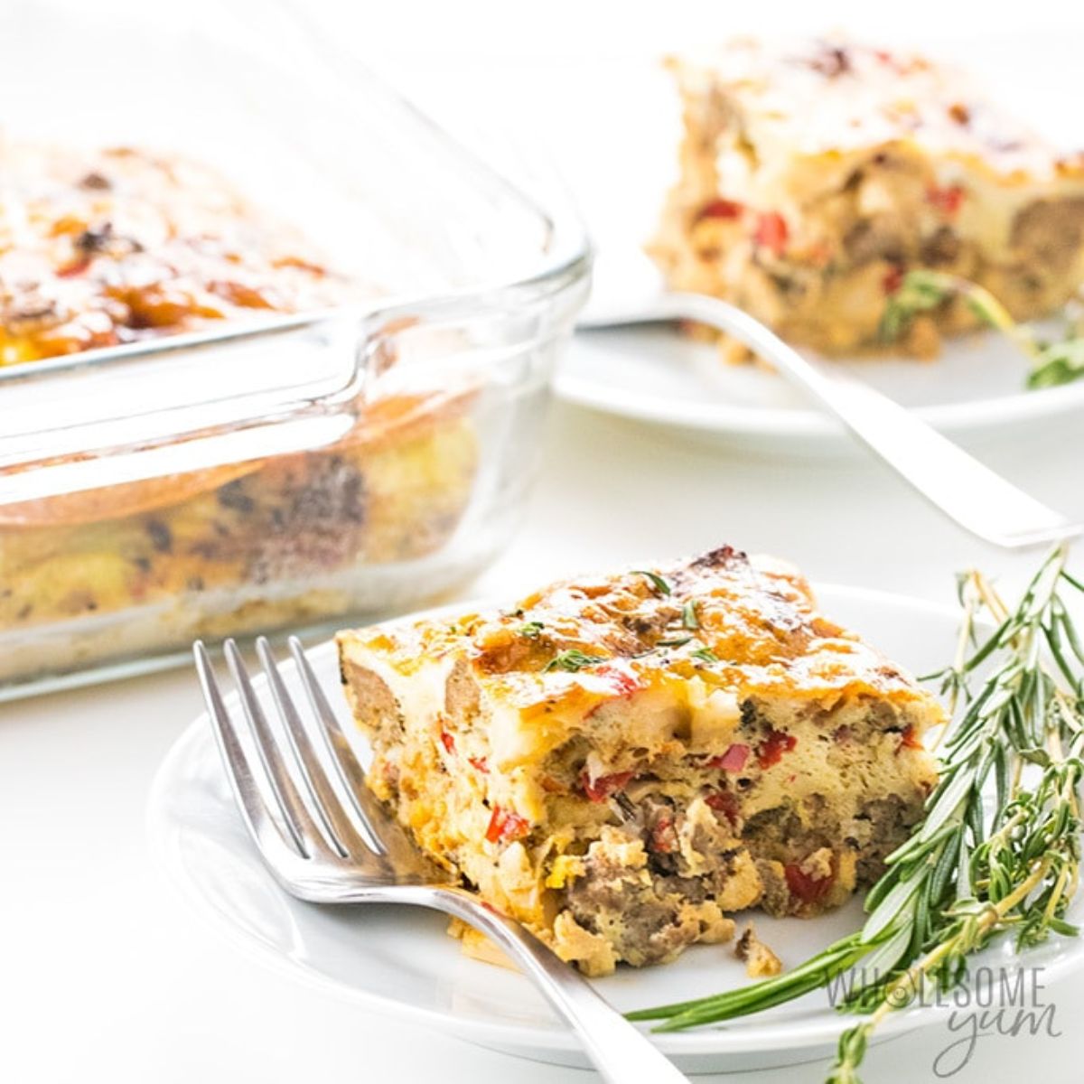 On a white tablecloth is a section of a glass casserole dish filled with casserole, and two plates of square portions of the casserole, one blurred.