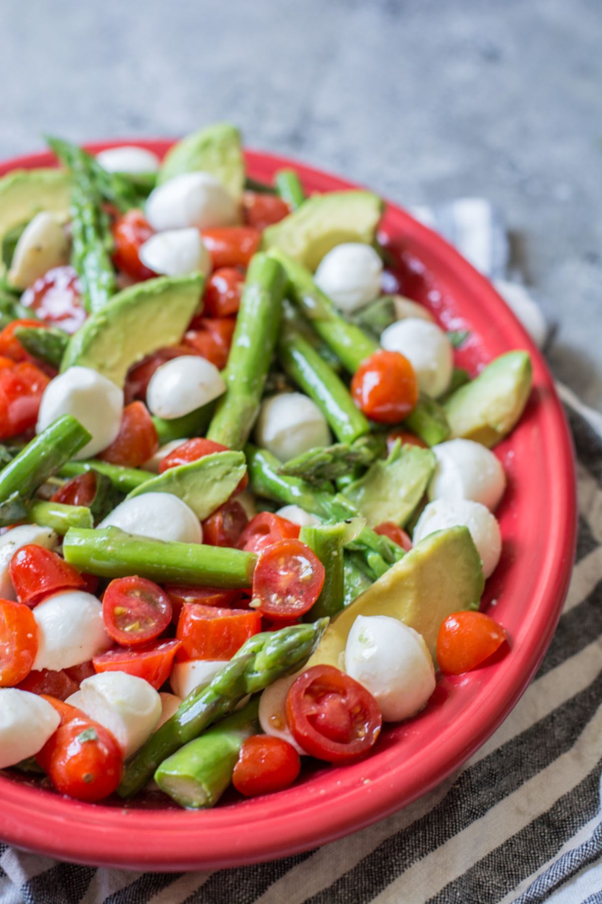 a red bowl sits on a blue and white striped cloth. In it is a salad made of asparagus stems, halved cherry tomatoes, mozzarella balls and avocado