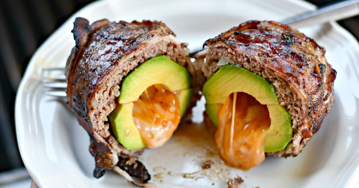 On a white plate are two halves of a avocado wrapped in hamburger meat and bacon, with a cheesy sauce oozing out of the middle. A silver fork is behind them