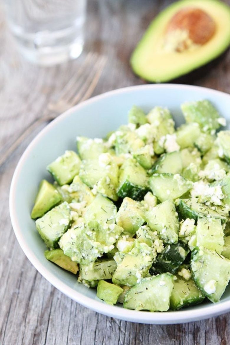 On a wooden table is a whote bowl with a cucumber, feta and avocado salad in it Behind is a lurred avocado half and the bottom of a glass