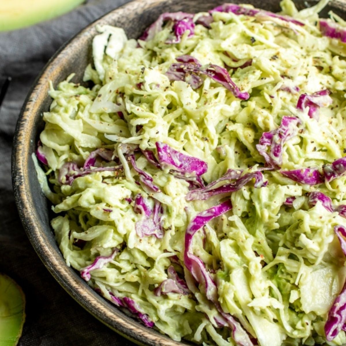 In a dark pottery bowl is a pile of coeslaw with red cabbage and other green sliced vegetables in a creamy sauce